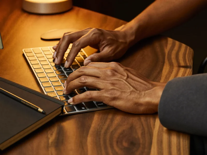 Hände auf beleuchteter Tastatur neben Notizbuch und Bleistift in warmem Licht